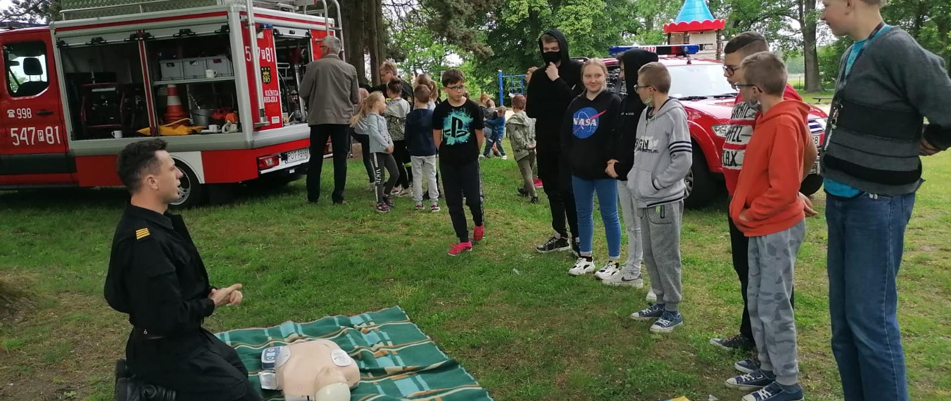 Zdjęcie przedstawia instruktaż pierwszej pomocy medycznej dla uczniów szkoły podstawowej. Na zdjęciu widzimy funkcjonariusza PSP, który prowadzi zajęcia związane z udzielaniem pierwszej pomocy oraz grupę dzieci uczestniczących w zajęciach. Na drugim planie widać samochód lekki, gdzie grupa dzieci jest zapoznawana ze sprzętem. Zdjęcie wykonane w porze dziennej na terenie szkoły.