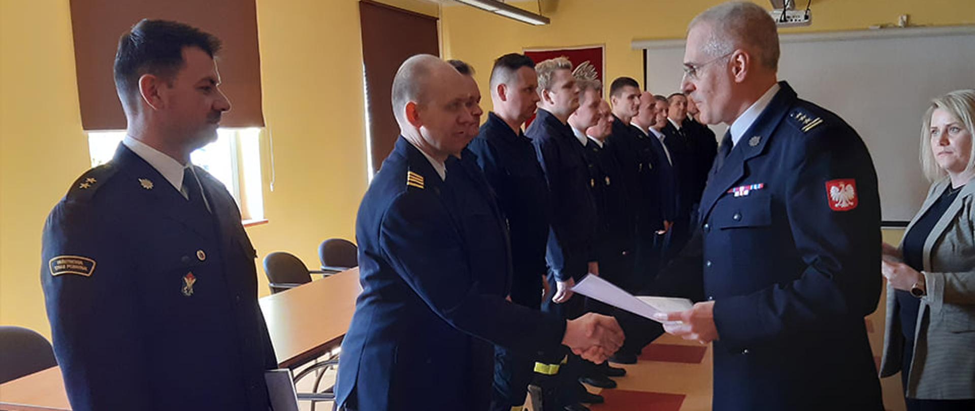 W Sali konferencyjnej w Komendzie Miejskiej Państwowej Straży Pożarnej w Wałbrzychu strażacy ubrani w ciemne mundury wyjściowe odbierają z rąk Komendanta nagrody i podziękowania za służbę
