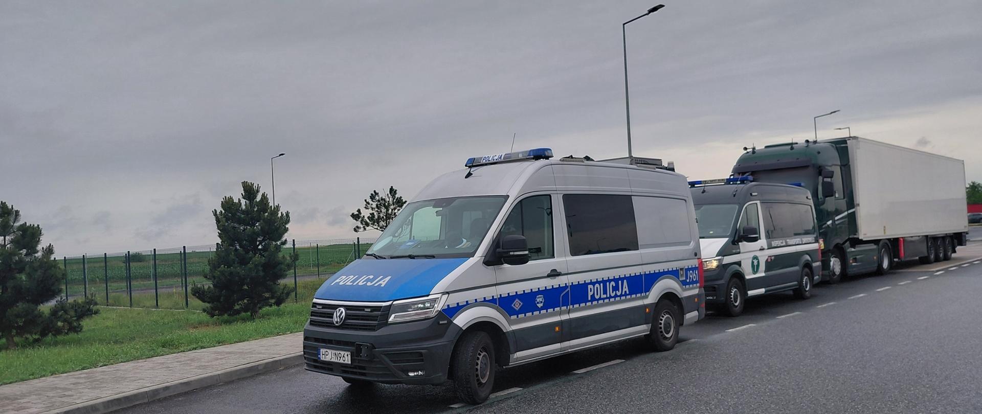 Srebrno-niebieski furgon policyjny marki Volkswagen oraz stojący za nim zielono-biały pojazd Inspekcji Transportu Drogowego, zaparkowane na poboczu drogi w pochmurny dzień. W tle widoczna jest zatrzymana zielona ciężarówka z białą naczepą oraz zielony pas zieleni z drzewami.