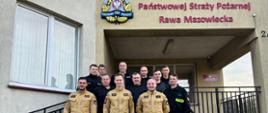 Grupa strażaków w mundurach służbowych i wyjściowych stoi przed wejściem do budynku Komendy Powiatowej Państwowej Straży Pożarnej w Rawie Mazowieckiej. Nad nimi widoczne jest logo oraz nazwa jednostki. 