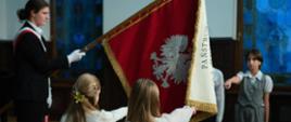 Uczniowie podczas ceremonii ślubowania w szkole, stoją przed sztandarem z godłem Polski