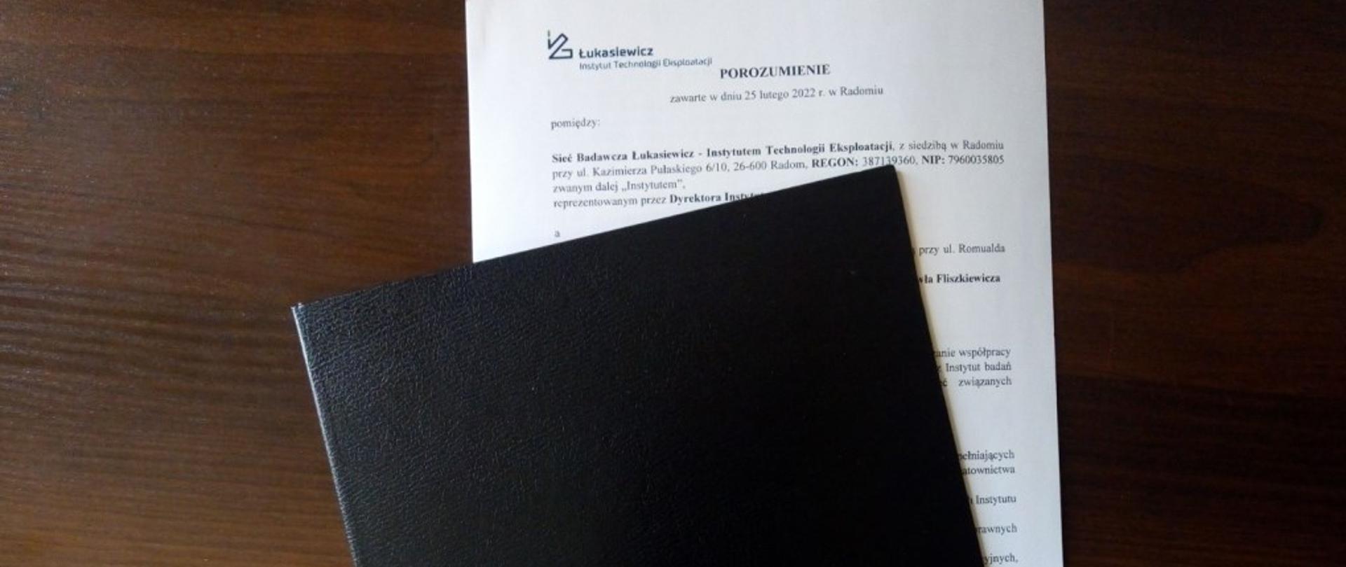 Zdjęcie przedstawia leżącą na brązowym biurku czarną teczkę do przenoszenia dokumentów. W prawym dolnym rogu teczki znajduje się biało - zielone logo Instytutu Technologii Eksploatacji w Radomiu. Z jej wnętrza wystaje kartka papieru w kolorze białym. Na kartce widać zapisaną treść porozumienia. 