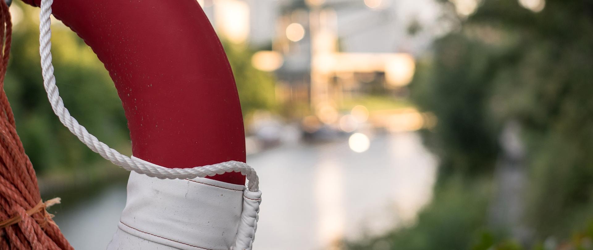 Zdjęcie przedstawia biało-czerwone koło ratunkowe, zawieszone na najpewniej moście. Pod nim w tle widać rzekę. Kadr ukazuje połowę koła, koło jest na pierwszym planie. Koło oplata gruby biały oraz pomarańczowy sznur. 