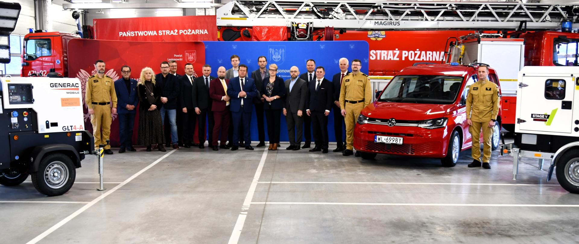 Na zdjęciu widać grupę kilkunastu osób stojących w garażu jednostki Państwowa Straż Pożarna. Są to zarówno strażacy w mundurach w kolorze piaskowym, jak i osoby w garniturach oraz stroju cywilnym, prawdopodobnie przedstawiciele władz lokalnych lub instytucji współpracujących.
W tle znajdują się czerwone wozy strażackie, w tym duży samochód z drabiną oraz czerwony samochód osobowy przystosowany do celów służbowych. Po bokach widoczne są także mobilne agregaty prądotwórcze na przyczepach. Za grupą ustawiono kolorowe ścianki z herbami i napisami, co sugeruje oficjalne wydarzenie, np. przekazanie sprzętu, uroczystość lub konferencję.
Wszyscy stoją w jednym rzędzie i patrzą w kierunku aparatu, co wskazuje, że jest to zdjęcie pamiątkowe z formalnej okazji.