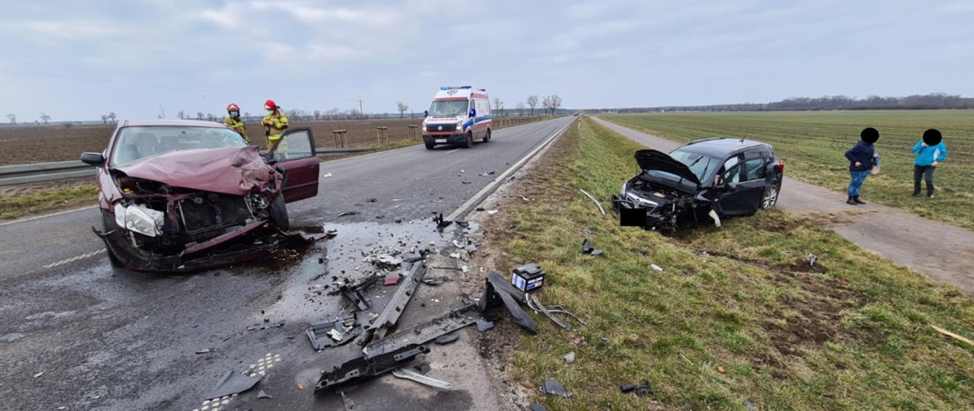 Widać dwa samochody, jeden na środku drogi z uszkodzonym przodem, drugi w rowie z uszkodzonym przodem, za nimi widać kartkę pogotowia