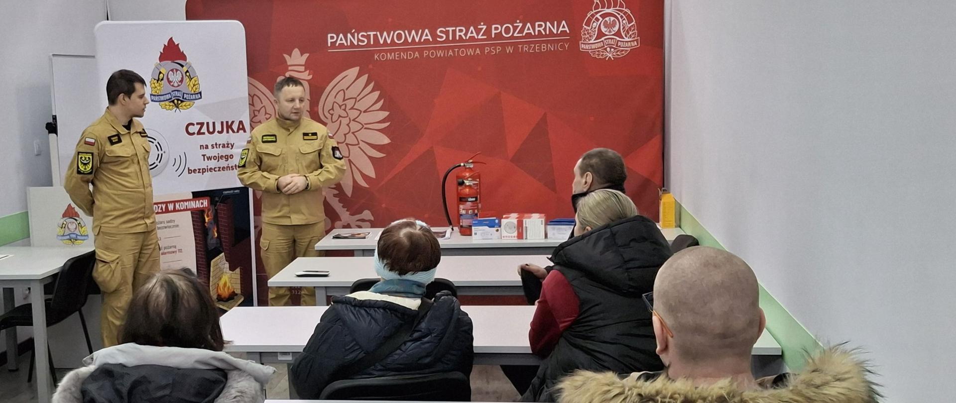 Zdjęcie przedstawia dwóch strażaków w jasnopiaskowych ubraniach specjalnych, którzy prowadzą prelekcję w sali szkoleniowej. Stoją oni na tle dużej, czerwonej ścianki promocyjnej z logo Państwowej Straży Pożarnej oraz pionowego baneru z napisem „CZUJKA”. Przed nimi, w ławkach szkolnych, siedzi tyłem do obiektywu grupa kilku słuchaczy w zimowych kurtkach, którzy uważnie obserwują i słuchają prowadzących.