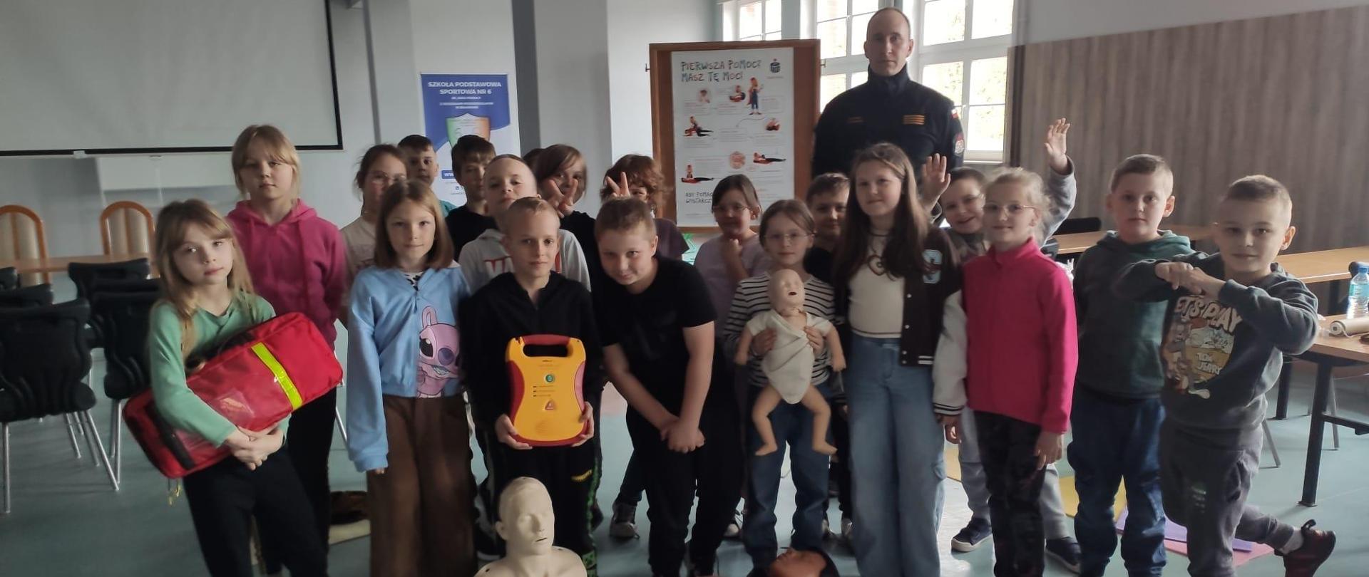 Na sali szkoleniowej stoi strażak z grupka dzieci. Pozują do zdjęcia ze szkoleniowym sprzętem medycznym, na podłodze leżą manekiny.