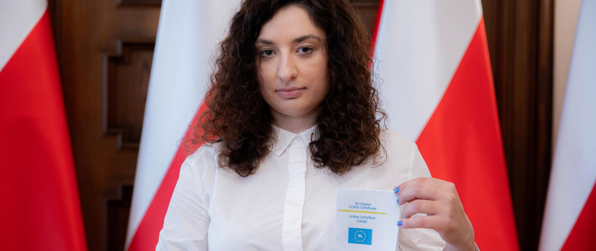 Pełnomocnik wojewody łódzkiego ds. szczepień przeciwko Covid-19 Jolanta Kowalik-Gęsiak stoi na tle flag w ŁUW i trzyma w ręce Unijny Certyfikat COVID