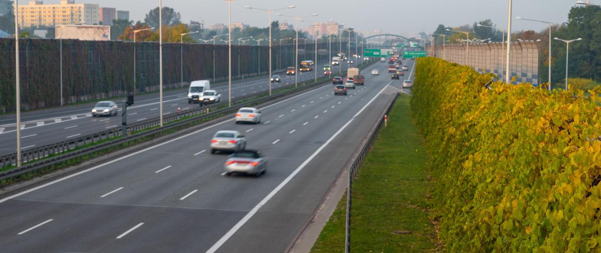 Pojazdy poruszające się drogą ekspresową. Pochmurny, jesienny dzień. Po obu stronach drogi stoją ekrany akustyczne. W oddali wiadukt.