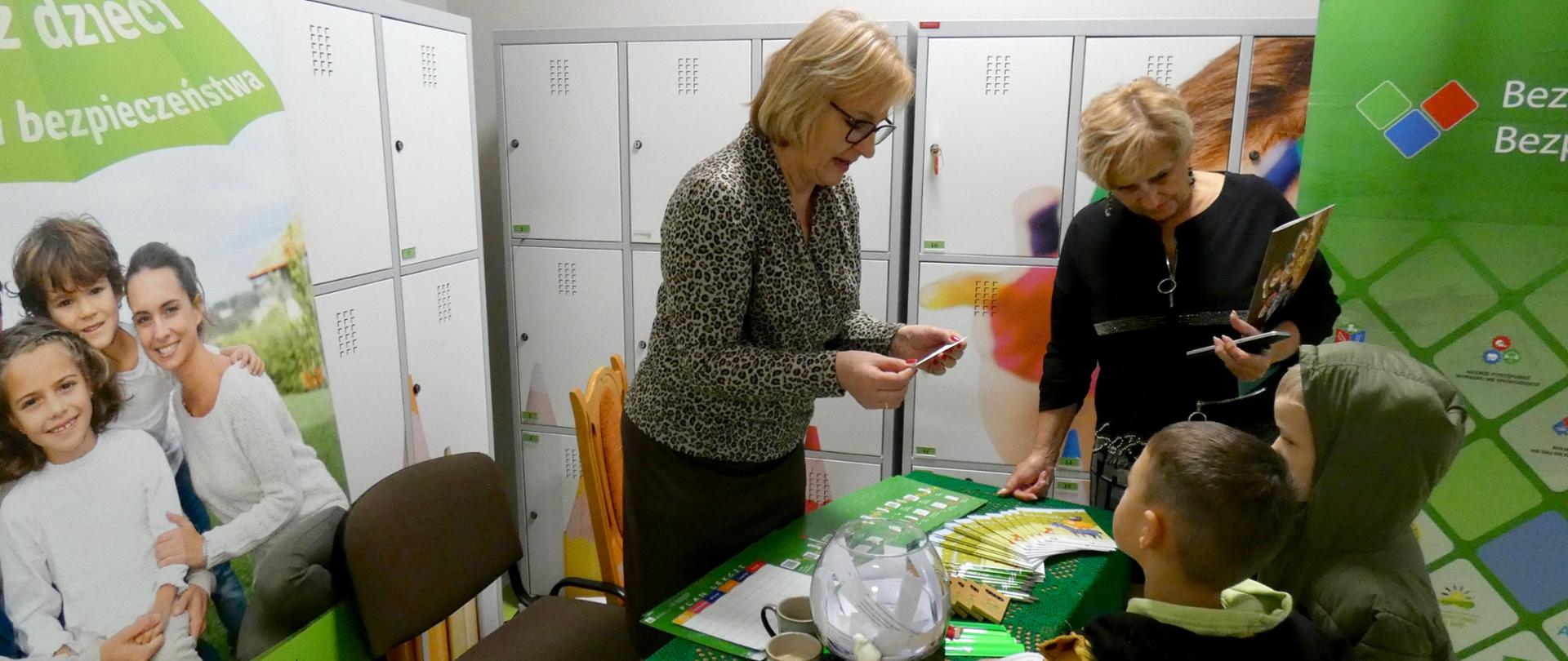 Pracownica KRUS i dyrektorka Szkoły Podstawowej prowadzą quiz o bezpieczeństwie w gospodarstwie rolnym z udziałem dwojga dzieci. Na stole rozłożone są broszury, kalendarze i materiały edukacyjne KRUS. W tle stoją banery z logo KRUS i hasłem „Bezpieczny Rolnik, Bezpieczna Wieś”.