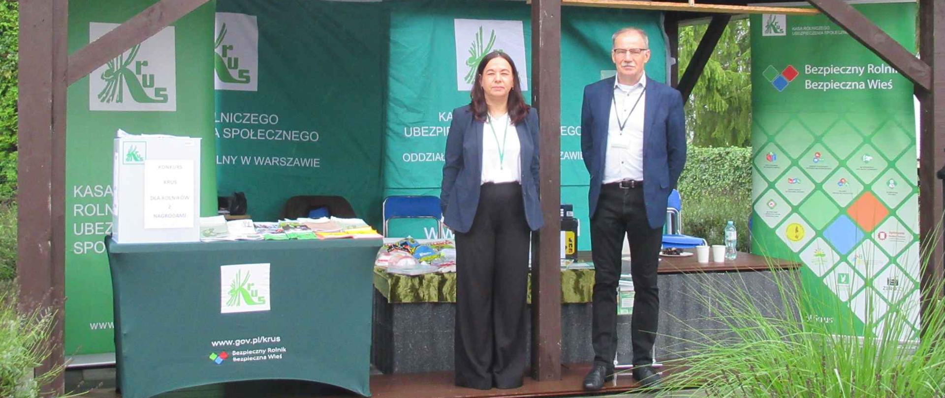 Pod drewnianą wiatą ustawiono stoisko PT KRUS z zielonymi banerami i stołem z materiałami informacyjnymi. Przed stoiskiem stoją dwie osoby, kobieta i mężczyzna – pracownicy KRUS.