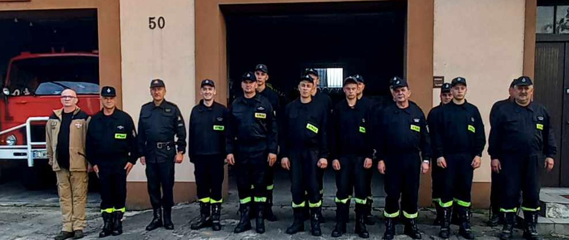 Przed budynkiem strażnicy w dwuszeregu stoją strażacy. Za nimi widać budynek strażnicy OSP Chociw z otwartymi bramami garażowymi. W jednej z bram widać czerwony samochód strażacki. Przed strażakami widać betonowy plac. 