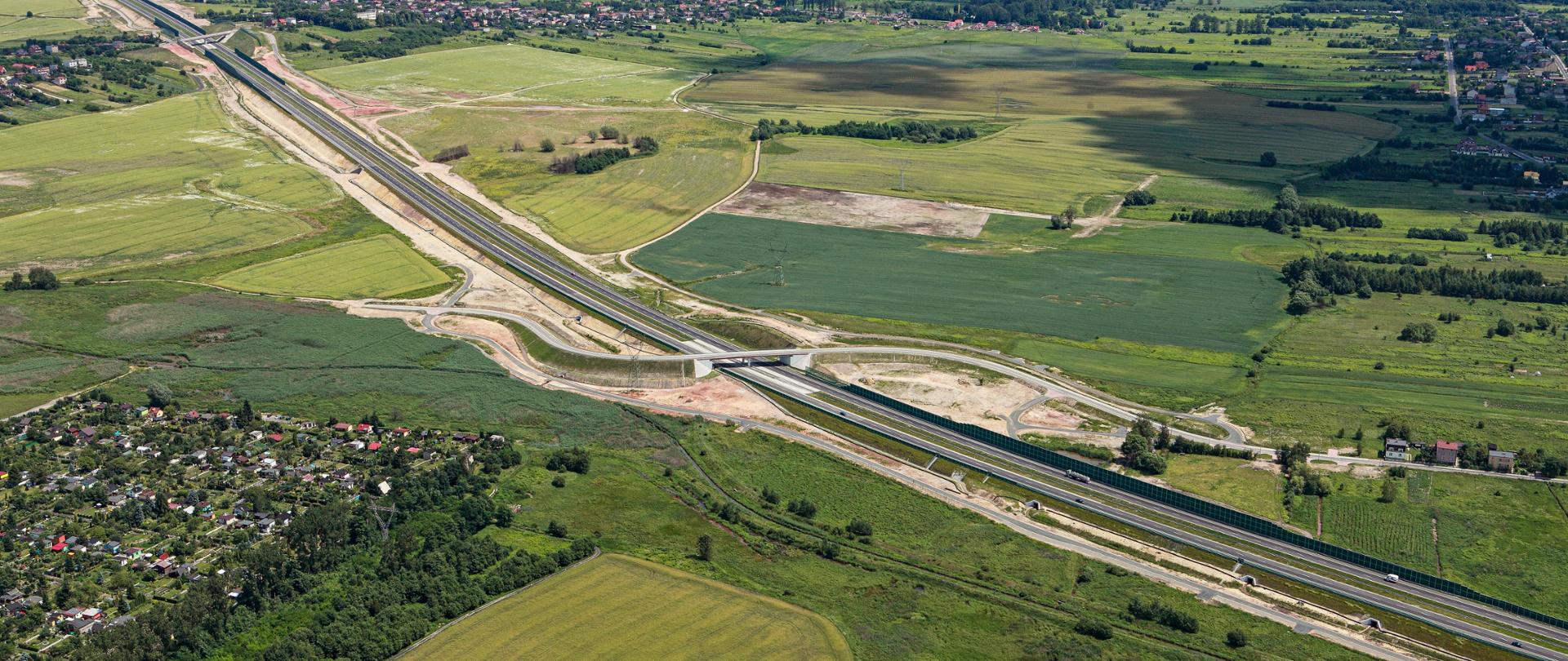 Fotografia lotnicza odcinka autostrady A1 pomiędzy Piekarami Śląskimi a Pyrzowicami. Jezdnia autostrady położona w południowej części na nasypie, w północnej w wykopie.  Pojedynczo przecinana  wiaduktami drogowymi nad autostradą. Otoczenia autostrady stanowią użytki rolne, w dalszej odległości zlokalizowane obszary zabudowy jednorodzinnej oraz leśne. Na fotografii dominują barwy jasno i ciemnozielone.  