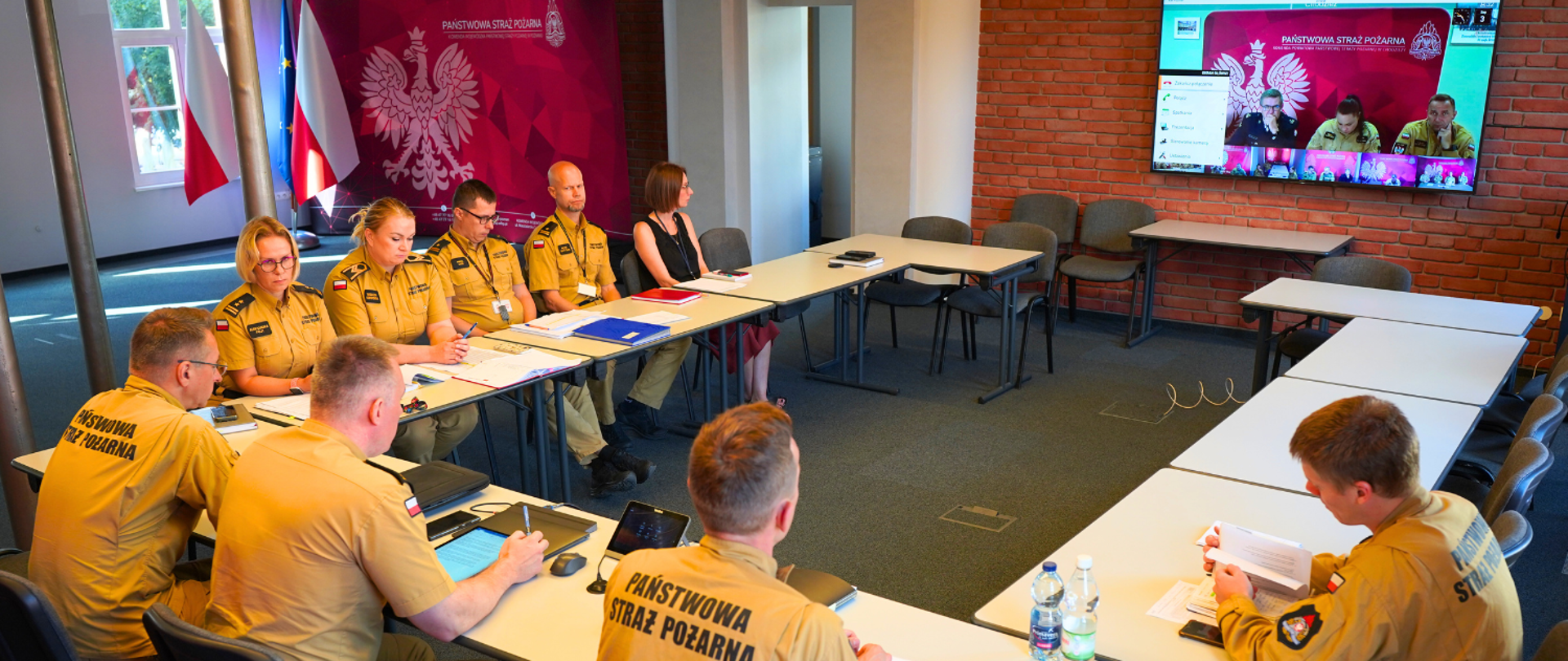 Panowie Komendanci, Państwo Naczelnicy oraz przedstawiciele wydziałów siedzą za stołami w sali komendy wojewódzkiej, uczestnicząc w wideokonferencji. Znajdują się przed telewizorem z transmisją. 