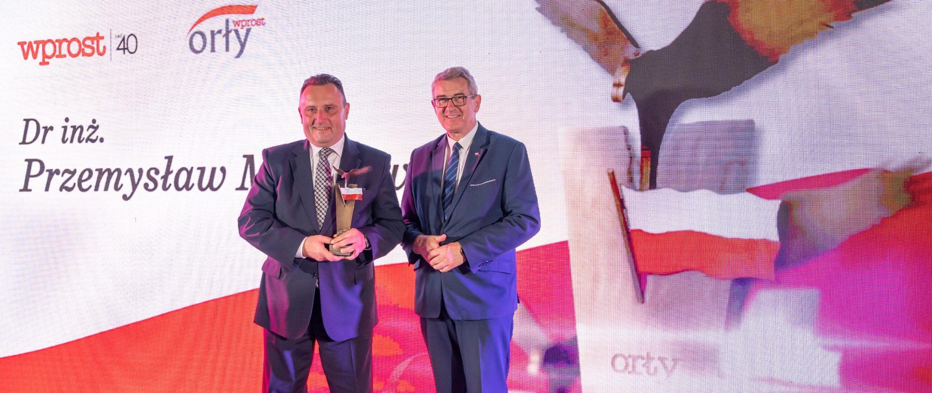 Wiceminister Murdzek stoi obok mężczyzny w garniturze który trzyma skrzydlatą statuetkę, za nimi ścianka z wizerunkiem wzlatującego orła.