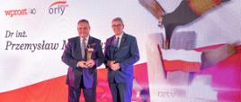 Wiceminister Murdzek stoi obok mężczyzny w garniturze który trzyma skrzydlatą statuetkę, za nimi ścianka z wizerunkiem wzlatującego orła.