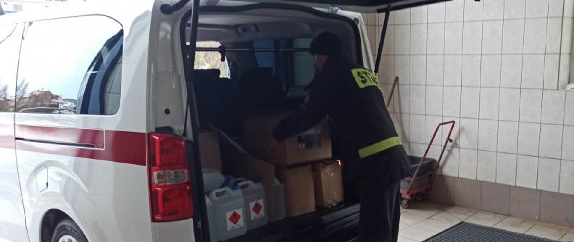 Teren komendy powiatowej PSP, budynek myjni, strażacy dokonują załadunku środków ochrony indywidualnej na samochód typu mikrobus