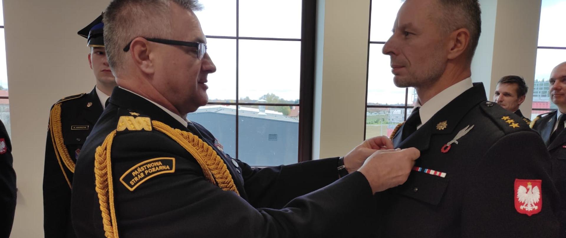 Nadbrygadier przypina medal funkcjonariuszowi. W tle widać kilku uczestników uroczystości. Wszyscy w mundurach galowych.