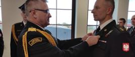 Nadbrygadier przypina medal funkcjonariuszowi. W tle widać kilku uczestników uroczystości. Wszyscy w mundurach galowych.