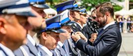 2. Małopolskie święto policji- wręczenie odznaczeń
Na pierwszym planie, w przybliżeniu, widoczny szereg funkcjonariuszy Policji oraz dwóch funkcjonariuszy Państwowej Straży Pożarnej. Naprzeciw nich Prezydent Miasta Krakowa wręcza odznaczenie „Honoris Gratia” – medal za zasługi dla miasta. W tle osoby uczestniczące w wydarzeniu oraz zabudowa krakowskiego Rynku Głównego.
