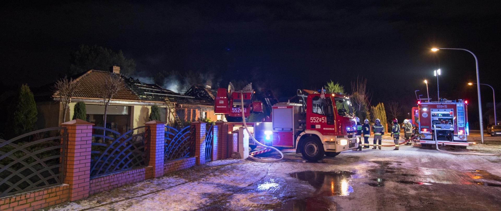 Na zdjęciu widać samochody pożarnicze, strażaków oraz w tle dach spalonego budynku.