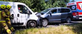 dwa samochody stoją przodem do siebie i mają zgniecione przody. obok stoi karetka