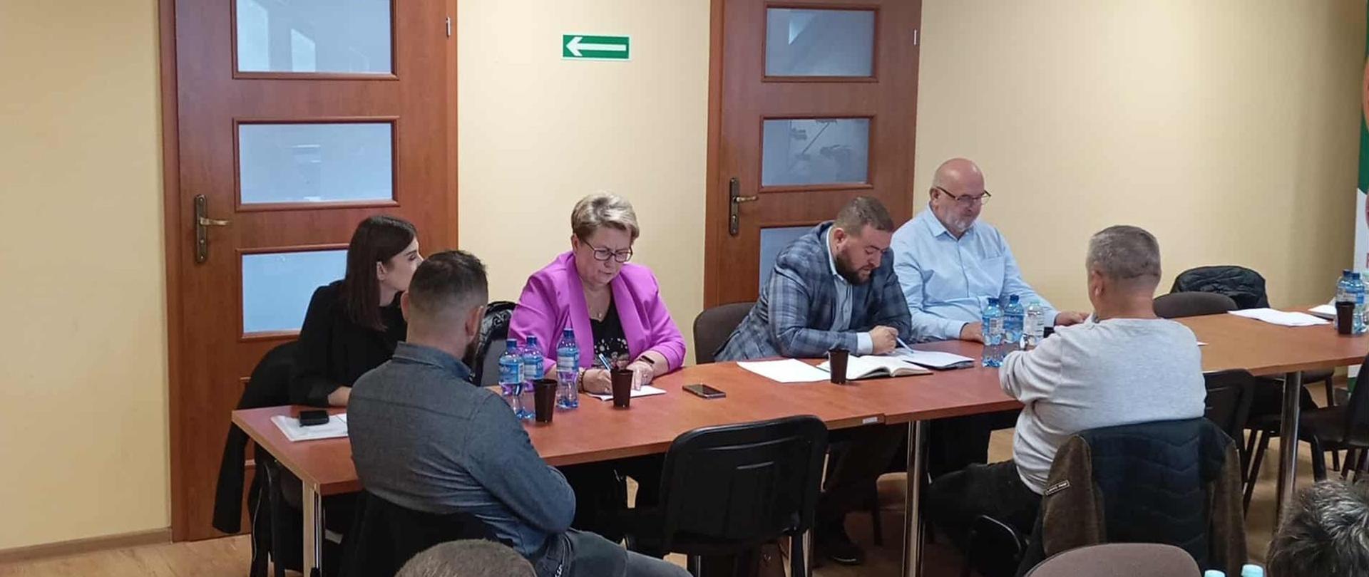 Kilka osób siedząca przy stołach. Na stołach porozmieszczane są dokumenty oraz butelki z wodą. Pomieszczenie jest jasne. Ściany są żółte.