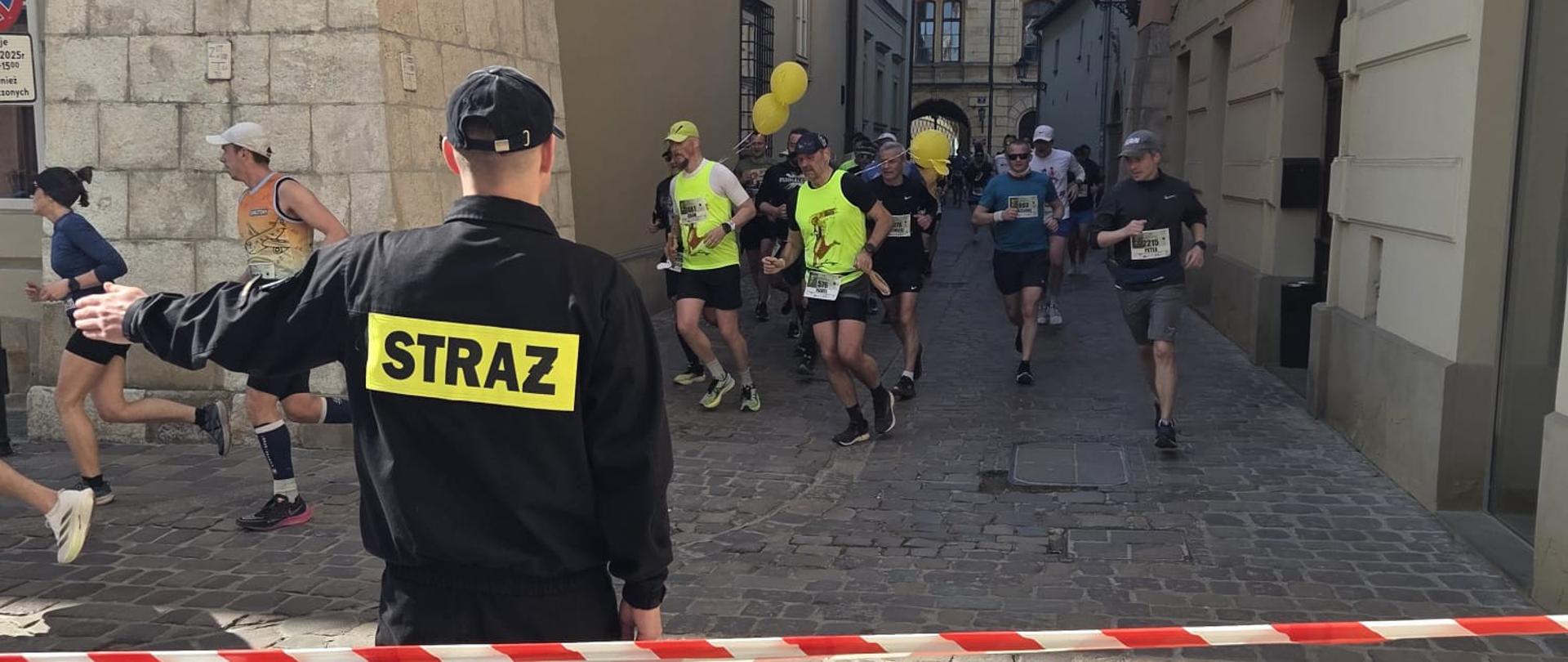 Zdjęcie przedstawia dynamiczny moment biegu „Krakowski Półmaraton Marzanny 2025”. Na pierwszym planie widać strażaka w czarnym ubraniu koszarowym z napisem straż na plecach. Stoi tyłem do obiektywu i wskazuje lewą ręką kierunek, biegnącym w tle osobom. W tle widoczna grupa biegaczy w sportowych strojach, która przemierza trasę wyścigu. Każdy z nich ma na sobie profesjonalny strój biegowy, numer startowy oraz sportowe okulary przeciwsłoneczne. Ich postawa i wyraz twarzy wskazują na wysiłek i koncentrację. Osoby znajdują się na wybrukowanej uliczce między budynkami o jasnych starych elewacjach. Na jednym z nich widnieje zielona tabliczka z nawą ulicy: Kanonicza. Słońce rzuca wyraźne cienie na chodnik, co sugeruje, że zdjęcie wykonano w pogodny dzień, a uczestnicy biegu rywalizują w doskonałych warunkach atmosferycznych.