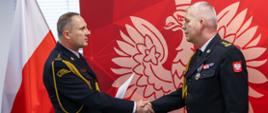 Dwóch wysokich rangą oficerów Państwowej Straży Pożarnej w galowych, czarnych mundurach podaje sobie dłonie podczas oficjalnej uroczystości. Stoją na tle czerwonej ścianki z godłem Polski, po lewej widoczna jest flaga biało-czerwona. Jeden z oficerów trzyma teczkę z dokumentami. Scena ma formalny, ceremonialny charakter.