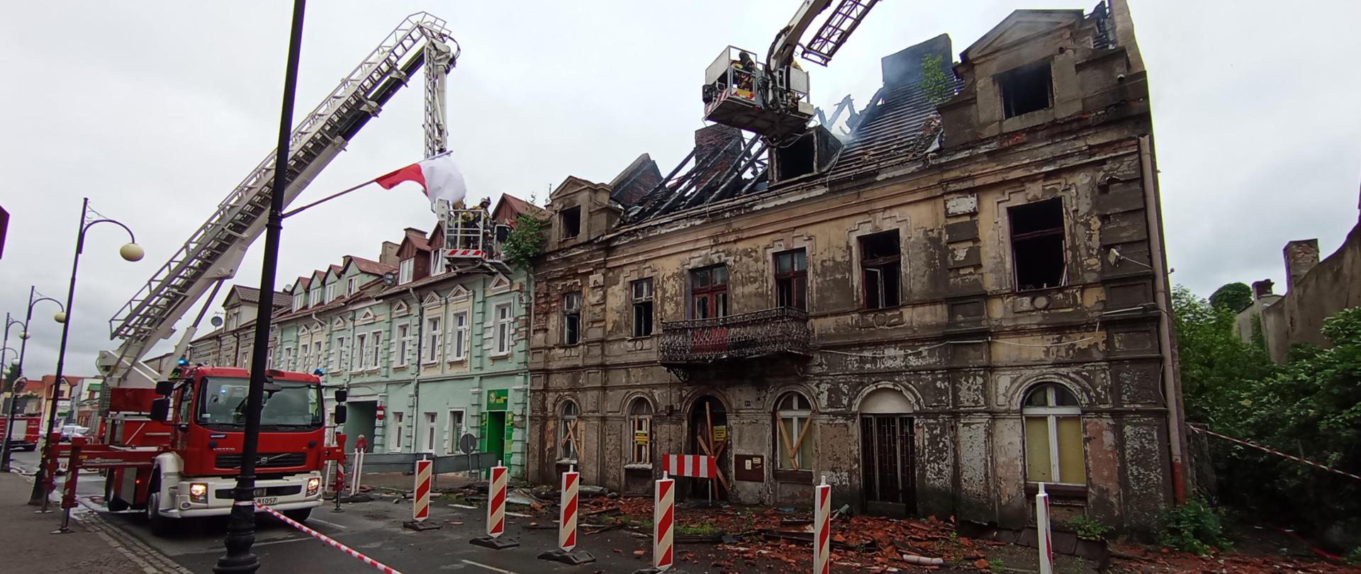 Zdjęcie przedstawia pożar kamienicy. Dach zawalony. Na chodniku widać gruz. Strażacy podają wodę z drabiny i podnośnika. 