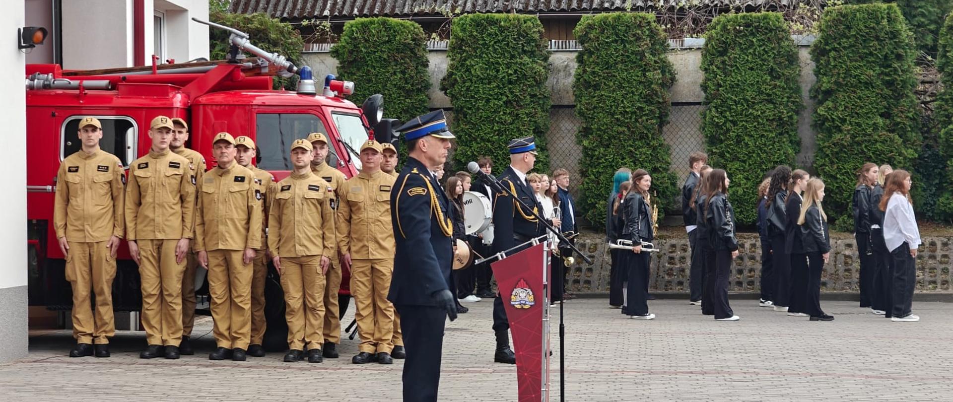 Fotografia przedstawia uroczystość plenerową przed strażnicą. Na pierwszym planie, przy nowoczesnej mównicy z mikrofonem, stoi oficer w ciemnym mundurze (granatowym lub czarnym) z pagonami i odznaczeniami, trzymający dokumenty. Obok niego stoi inny umundurowany mężczyzna. Po lewej stronie, wzdłuż zabytkowego, czerwonego wozu strażackiego, stoi szpaler mężczyzn w beżowych kombinezonach ochronnych, stojących na baczność. Na budynku po lewej widać dużą polską flagę (biało-czerwoną) zamontowaną na uchwycie ściennym. Po prawej stronie, na brukowanym placu, stoi spora grupa młodzieży i obserwatorów, głównie w ciemnych kurtkach i spodniach, ustawionych w rzędach. Niektórzy z nich wydają się mieć instrumenty muzyczne. Tło stanowią budynki o różnym charakterze oraz rząd wysokich, kolumnowych tuj. Na placu widać również fragmenty nawierzchni z kostki brukowej i klinkierowej.