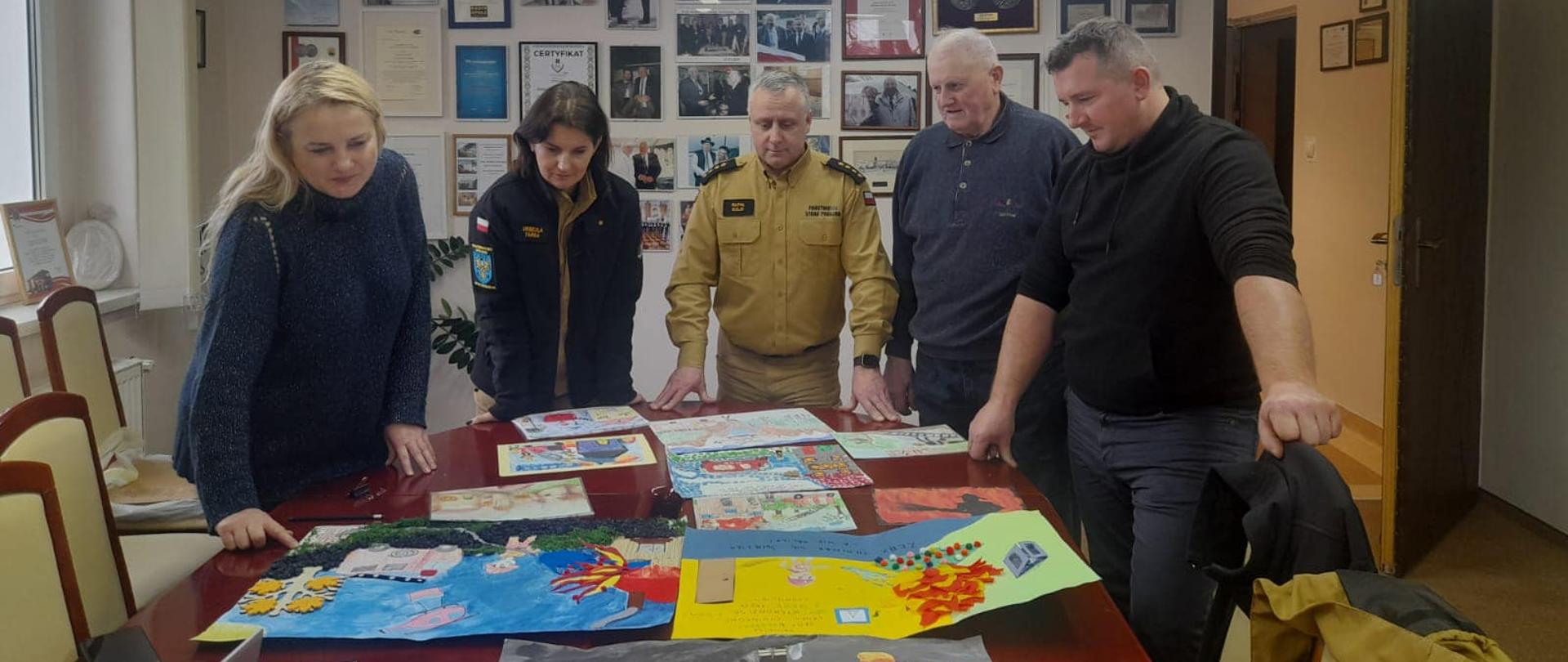 Na zdjęciu znajduje się jury (dwie kobiety i trzech mężczyzn) które stoi przy stole na którym znajdują się prace plastyczne