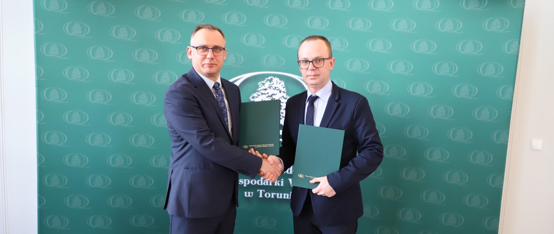 Sygnatariusze umowy przy stole, za nimi zielony baner z napisem WFOŚiGW