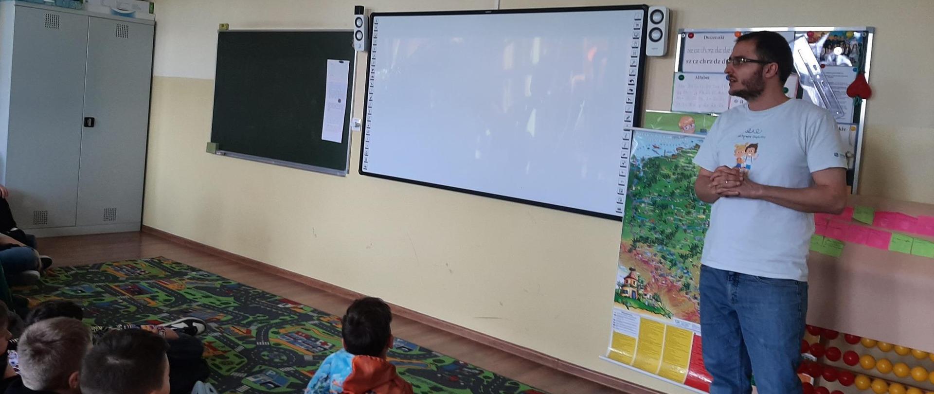 Szkoła Podstawowa w Kruchowie. Klasa w szkole. Zdjęcie zrobione z tyłu klasy. Uczniowie siedzą na dywanie. Naprzeciw nich wykładowca. Mówi. W tle prezentacja. 