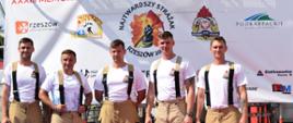 Na zdjęciu widzimy stojących w szeregu pięciu strażaków ubranych w białe koszulki i spodnie nomex. w tle widoczny jest baner tytułowy memoriału.