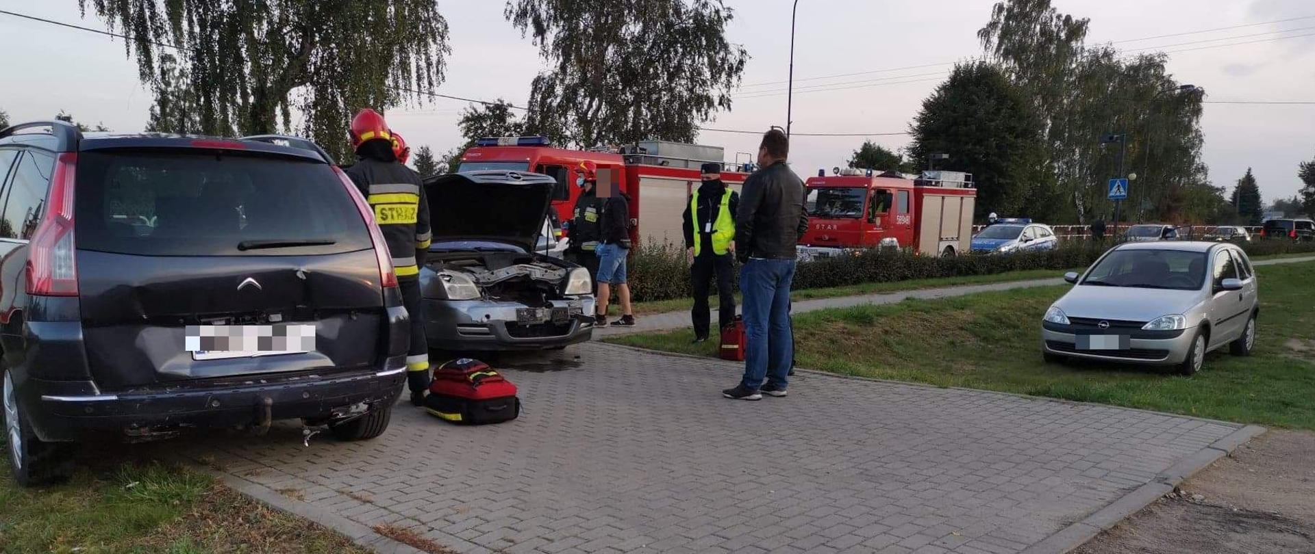 Na zjeździe z drogi stoją dwa samochody. Jeden przodem z podniesiona maską, drugi tyłem. Strażacy i policjanci prowadzą akcję. Na ziemi leży torba medyczna. W tle stoją wozy strażackie oraz radiowóz policyjny. 