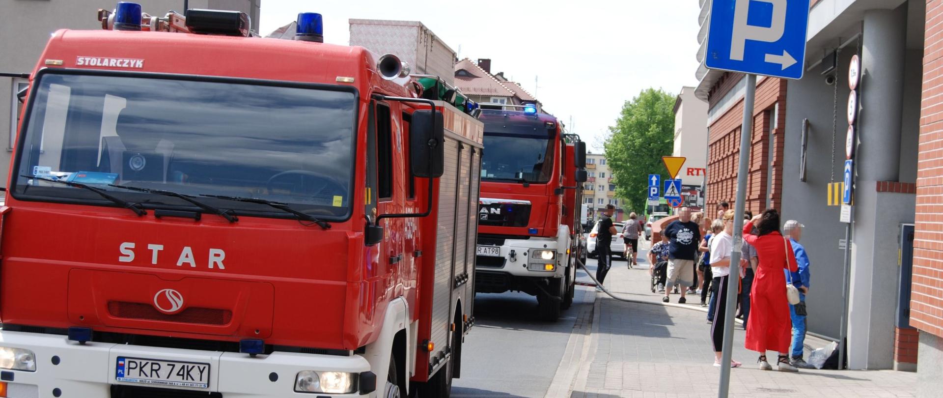Zdjęcie przedstawia samochody pożarnicze zaparkowane przed wjazdem do podziemnego parkingu galerii handlowej. Na chodniku tłum ludzi, którzy opuścili galerię.
