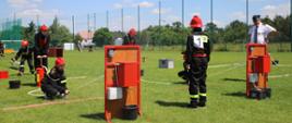 Na zdjęciu młodzieżowa drużyna pożarnicza w czarnych strojach i czerwonych hełmach nalewa wodę do nalewaków przy pomocy hydronetek.