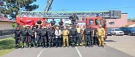 Wspólne zdjęcie grupowe uczestników szkolenia na drabiny mechaniczne. Strażacy w umundurowaniu służbowym i koszarowym. W tle stoją dwie drabiny mechaniczne należące do jednostek OSP.