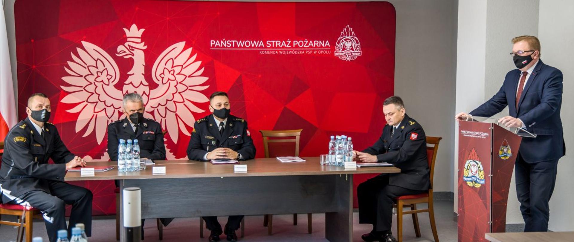 Przy mównicy przemawia Wicewojewoda Opolski Pan Tomasz Witkowski. Przy stole prezydialnym siedzą: Opolski Komendant Wojewódzki PSP w Opolu st. bryg. Krzysztof Kędryk, st. bryg. Wojciech Nawara Zastępca OKW PSP w Opolu, st. bryg. Arkadiusz Kuśmierski Zastępca OKW PSP w Opolu oraz dh Andrzej Borowski Prezes Oddziału Wojewódzkiego Związku Ochotniczych Straży Pożarnych Rzeczypospolitej Polskiej Województwa Opolskiego. W tle widoczna czerwona ścianka z białym napisem Państwowa Straż Pożarna i Komenda Wojewódzka PSP w Opolu. Na ściance białe godło Polski.