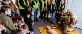 Strażak w namiocie pneumatycznym instruuje dzieci jak prawidłowo wykonywać resuscytację krążeniowo-oddechową na fantomie szkoleniowym, w tle dzieci obserwujące pokaz