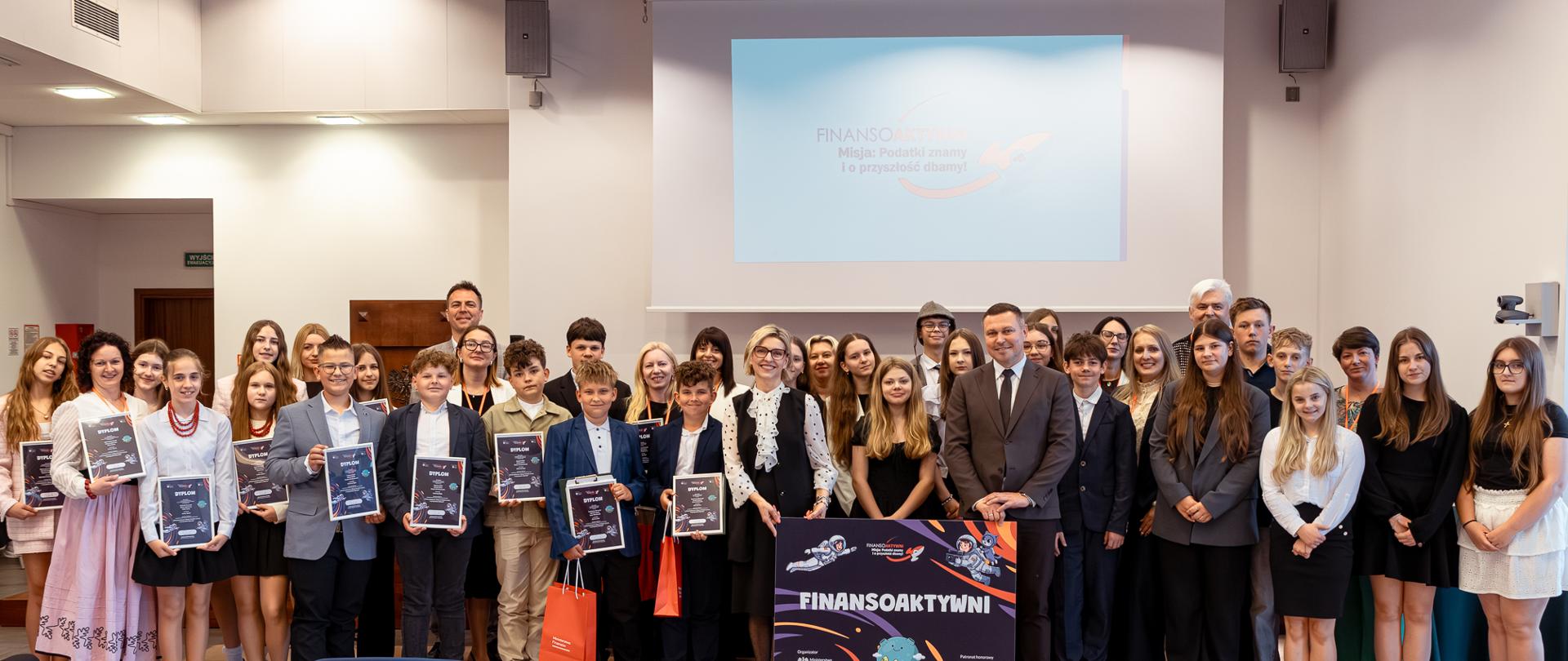Grupa osób stoi w sali konferencyjnej, część trzyma dyplomy, a na środku widoczny jest plakat „FINANSOAKTYWNI” przed ekranem projekcyjnym.
