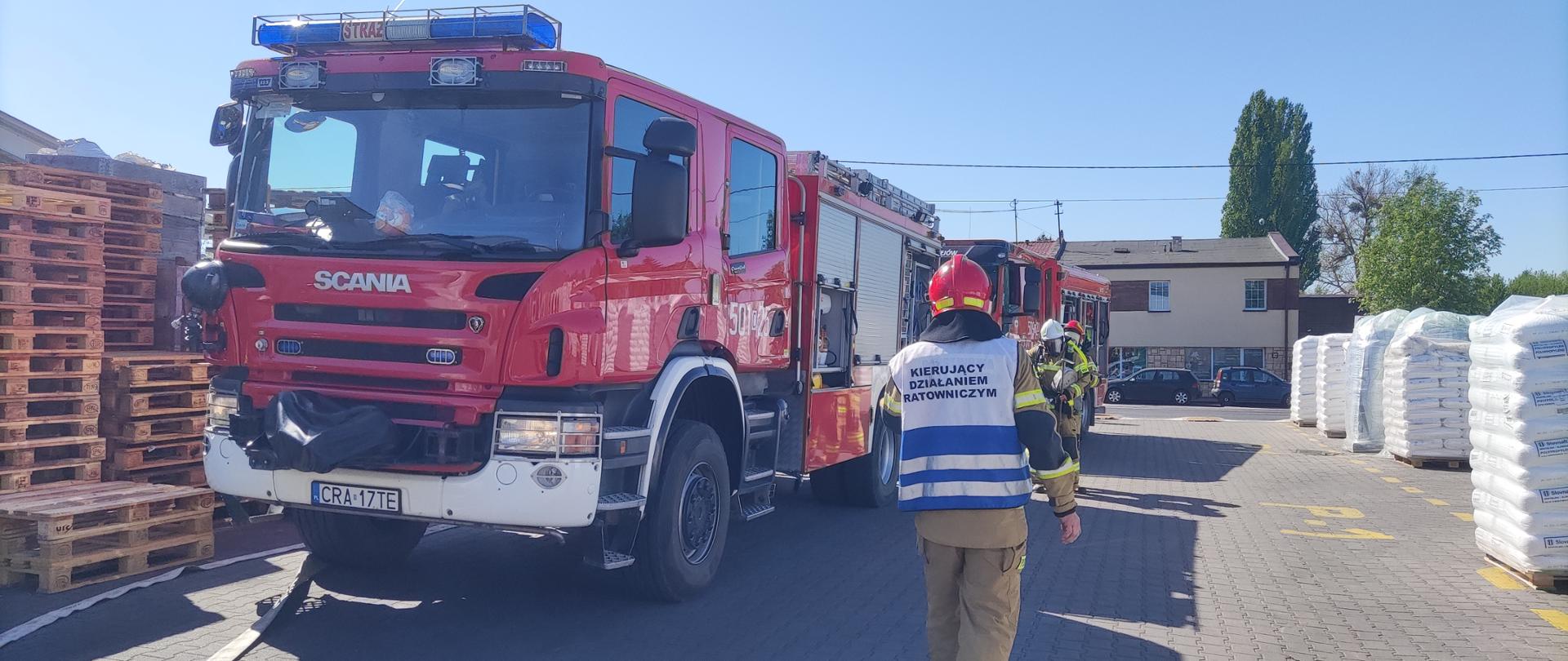 Zdjęcie przedstawia teren zakładu, w którym prowadzone były ćwiczenia obiektowe, na pierwszym planie dwa wozy strażackie oraz strażacy podczas ćwiczeń.