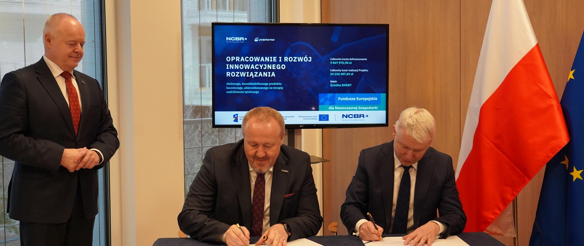 Przy stoliku siedzi dwóch mężczyzn w garniturach, podpisują leżące dokumenty, za nimi stoi na stojaku monitor z napisem Opracowanie i rozwój innowacyjnego rozwiązania, z lewej nad stolikiem stoi wiceminister Gzik, z prawej pod ścianą flagi Polski i UE.