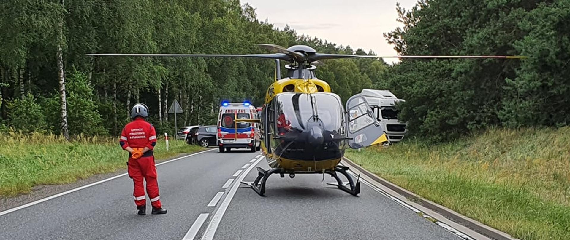 Na fotografii znajduje się śmigłowiec Lotniczego Pogotowia Ratunkowego, który wylądował na środku drogi. Śmigłowiec jest koloru żółtego. Dookoła znajduje się trawa oraz drzewa. Po lewej stronie znajduje się pilot w czerwonym kombinezonie oraz siwym hełmie. Za śmigłowcem po lewej stronie fotografii znajduje się karetka pogotowia oraz dwa samochody osobowe. Po prawej stronie znajduje się biały ciągnik siodłowy. Zdjęcie wykonano w porze dziennej.