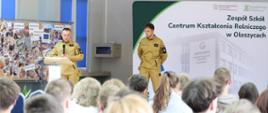 Na scenie stoją dwaj druhowie z Ochotniczej Straży Pożarnej w Oleszycach ubrani w piaskowe mundury wyjściowe. Jeden ze strażaków przemawia do zgromadzonych przez mikrofon przy mównicy, podczas gdy drugi z powagą asystuje mu obok oficjalnej ścianki z logo szkoły.