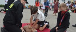 Boisko Zespołu Szkolno- Przedszkolnego nr 2 w Rawiczu. Strażak, członek Ochotniczej Straży Pożarnej w Sarnowie uczy dzieci udzielania pierwszej pomocy. Chłopiec klęczy przy fantomie i uciska klatkę piersiową. Obok niego dwoje innych uczniów. W tle grupa uczniów przyglądająca się pokazowi.