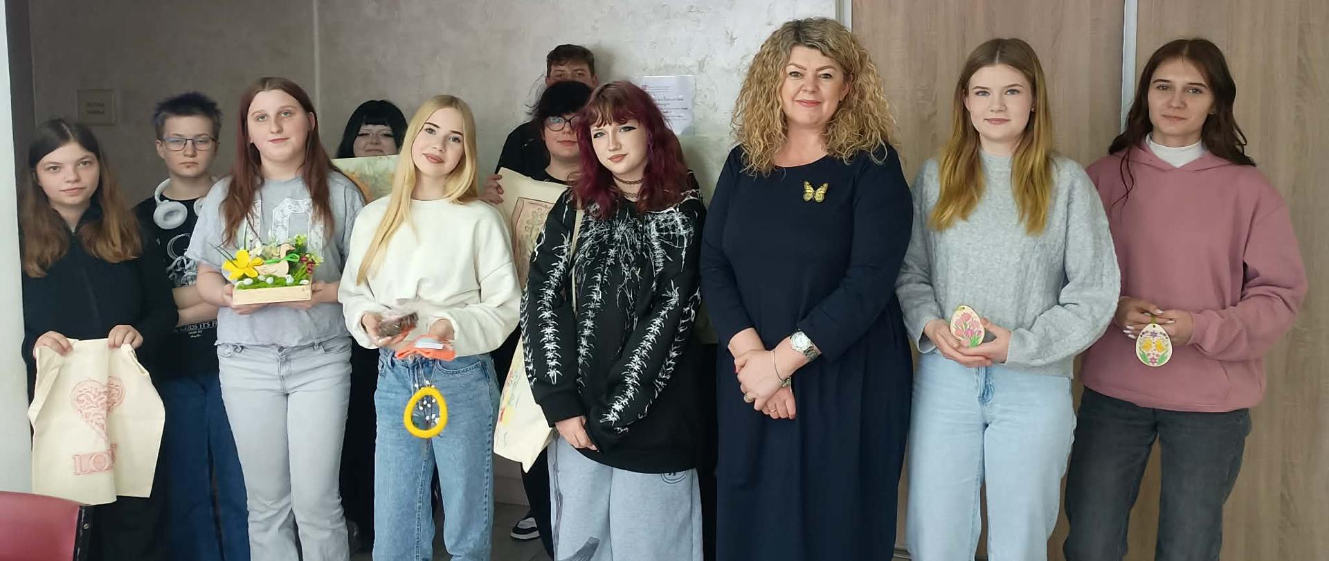 Grupa uczniów wraz z Kierownikiem Warsztatu Terapii Zajęciowej (WTZ) w Oleszycach stoi w pomieszczeniu warsztatowym. Uczniowie trzymają własnoręcznie wykonane prace, m.in. ozdobione torby, stroiki świąteczne i dekoracje. W tle widoczne są drzwi oraz godło Polski.