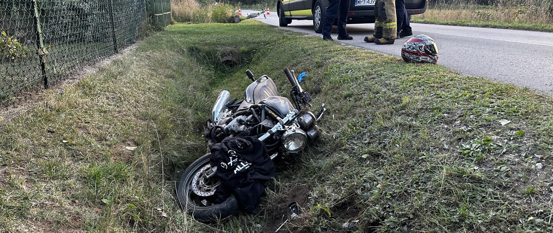 Zdjęcie przedstawia miejsce wypadku drogowego. Na poboczu drogi, w płytkim rowie porośniętym trawą, leży przewrócony motocykl. Obok motocykla leży ciemna kurtka. Na asfalcie, obok trawy, leży kask motocyklowy. Dalej na drodze widoczna jest karetka pogotowia ratunkowego, a obok niej stoją osoby w odblaskowych kamizelkach i mundurach służb ratunkowych. Po lewej stronie zdjęcia widać zielone ogrodzenie. Pogoda jest pochmurna, ale jest jasno.