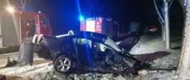 Na zdjęciu widoczny jest uszkodzony samochód osobowy stojący na kołach w rowie, na drodze znajdują się pojazdy strażackie